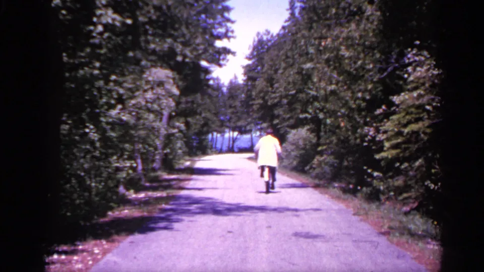 The cyclist rides away from the camera along the quiet paved road, surrounded by dense greenery in Michigan home-movie footage.