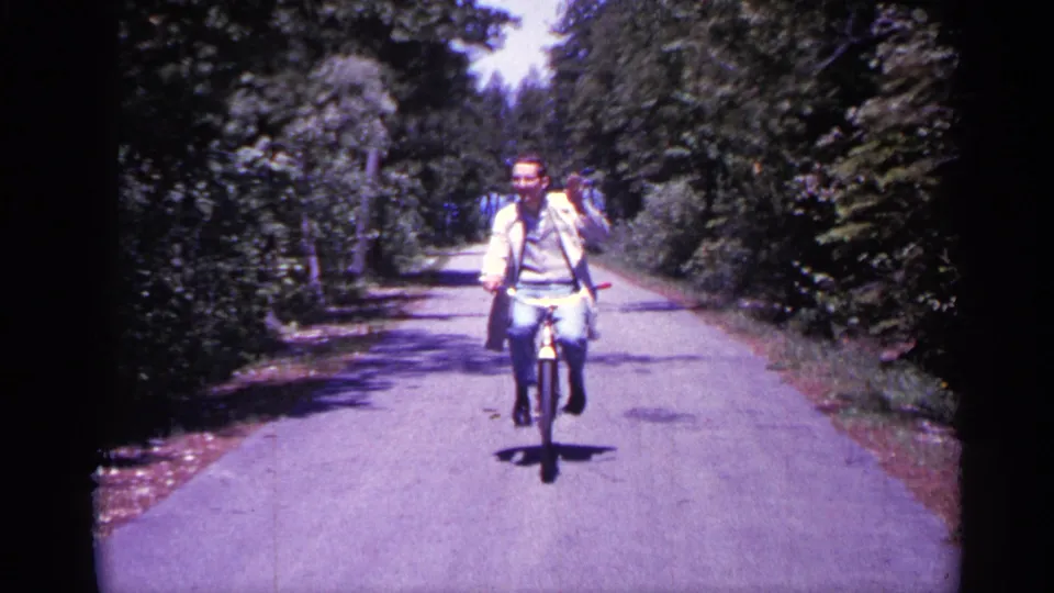 A smiling adult man in glasses rides closer to the camera on a quiet Michigan road in 1960 home-movie footage.