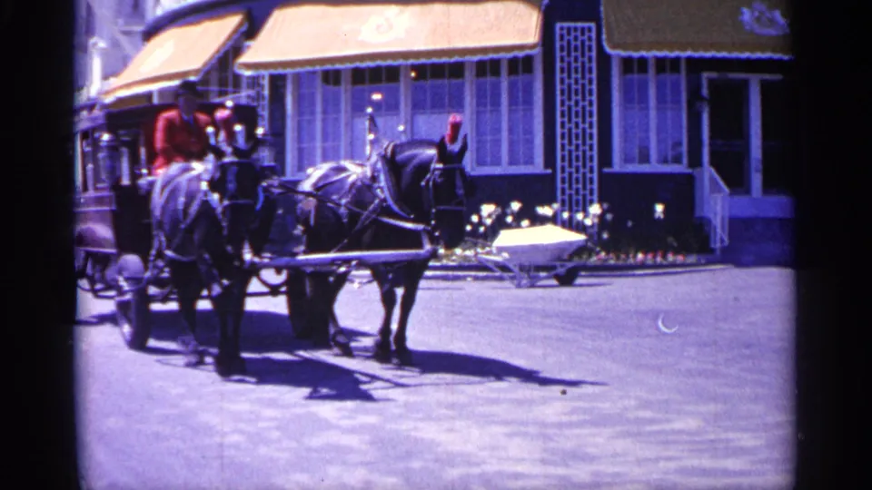 1960 Michigan home-movie frame showing a man in a red jacket driving a horse-drawn carriage past a storefront with a yellow awning.