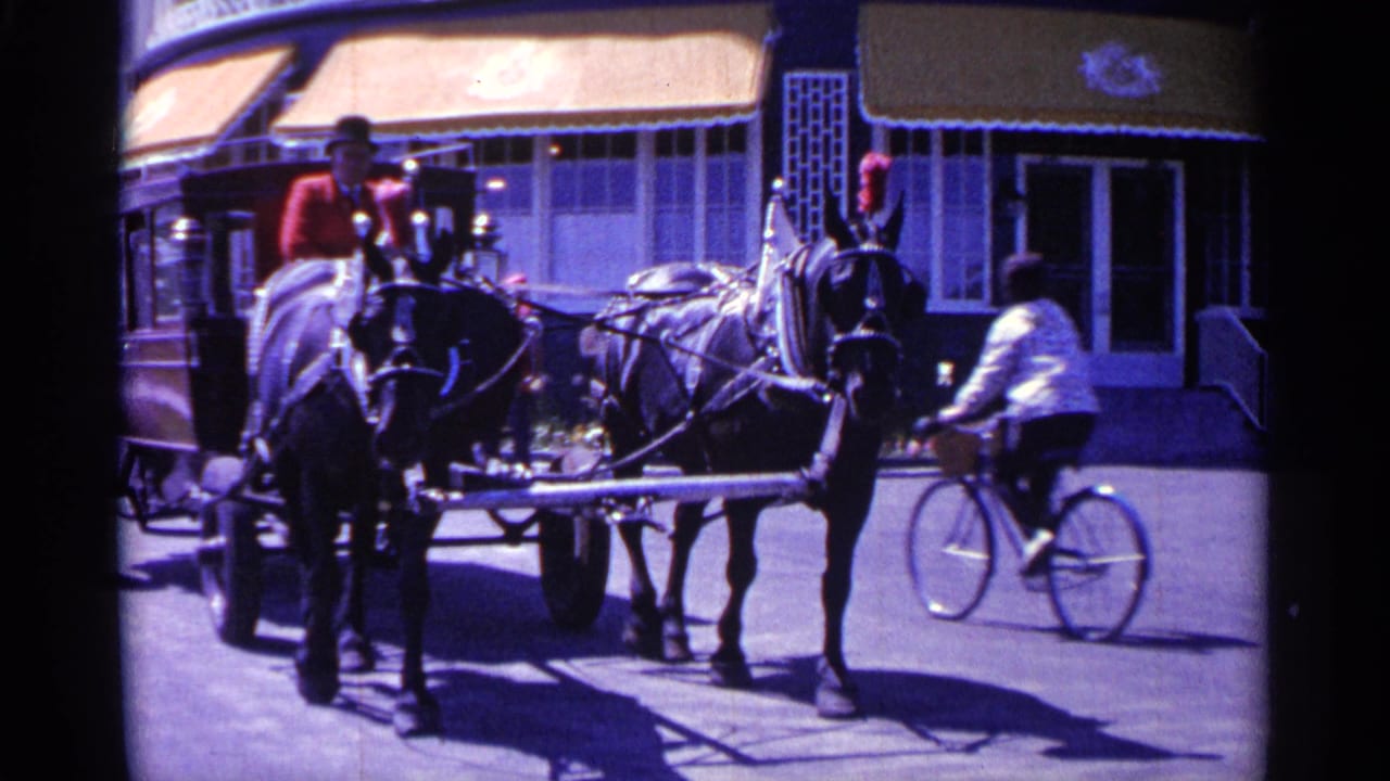 Two harnessed horses and a red cart stand near storefronts while a cyclist passes behind them.