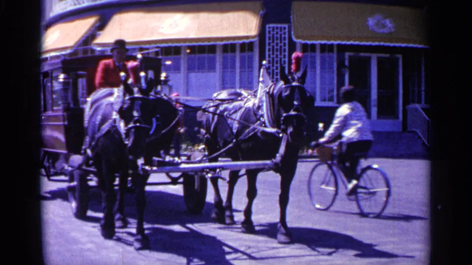 1960 Michigan 8mm frame showing two harnessed horses with a red passenger cart near storefront awnings as a cyclist rides past.