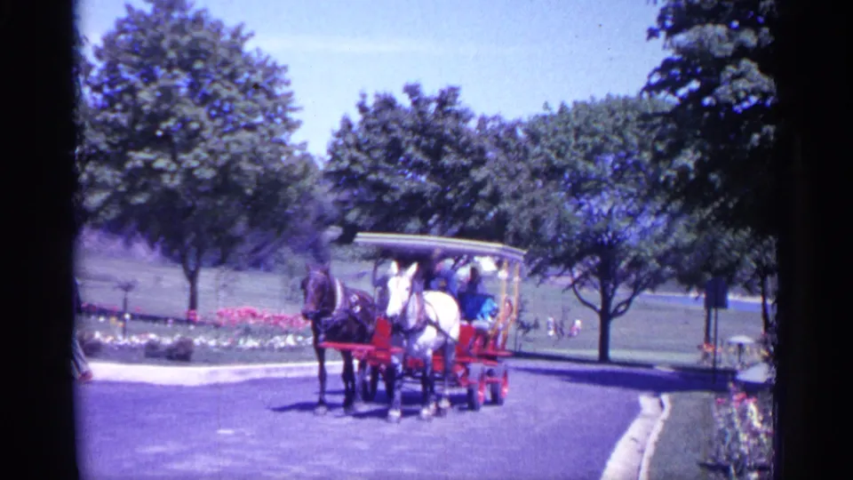1960 Michigan 8mm frame of a red horse-drawn passenger cart with a white canopy moving through a landscaped park road.