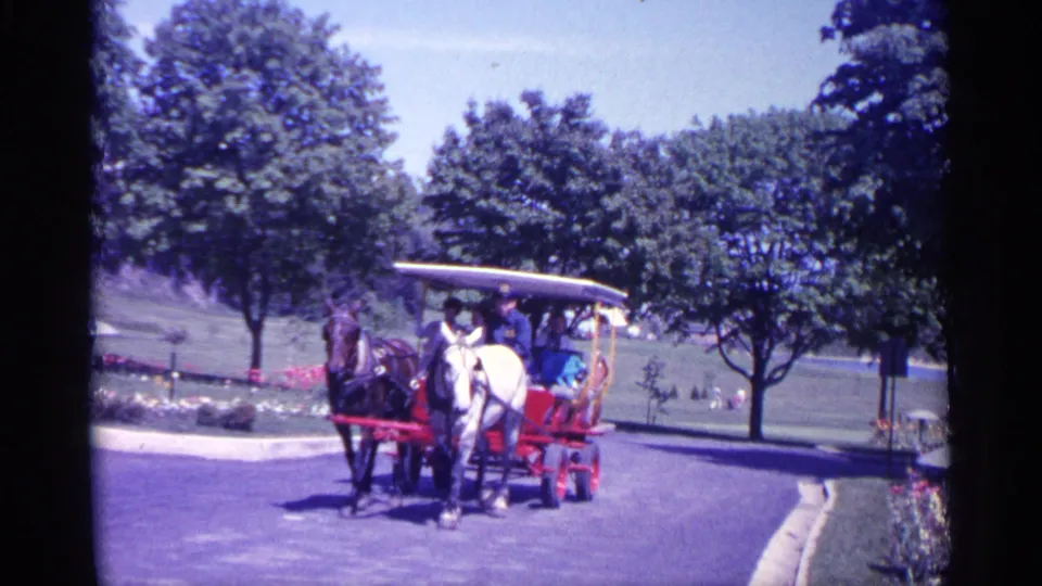 1960 Michigan 8mm frame showing a dark horse and white horse pulling a red passenger cart through a green park setting.