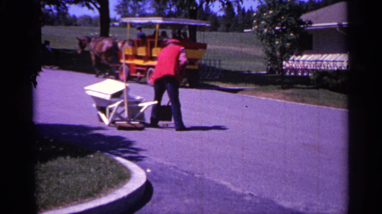 Worker with broom and wheelbarrow near a carriage pulled by horses on a paved road.