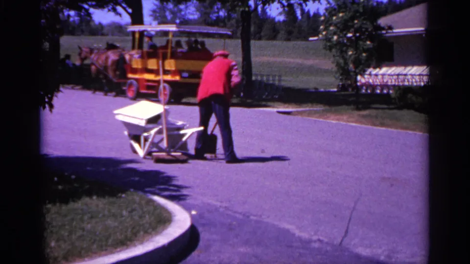 A mid-century home-movie view shows the carriage and horses passing behind a worker sweeping beside a white wheelbarrow.