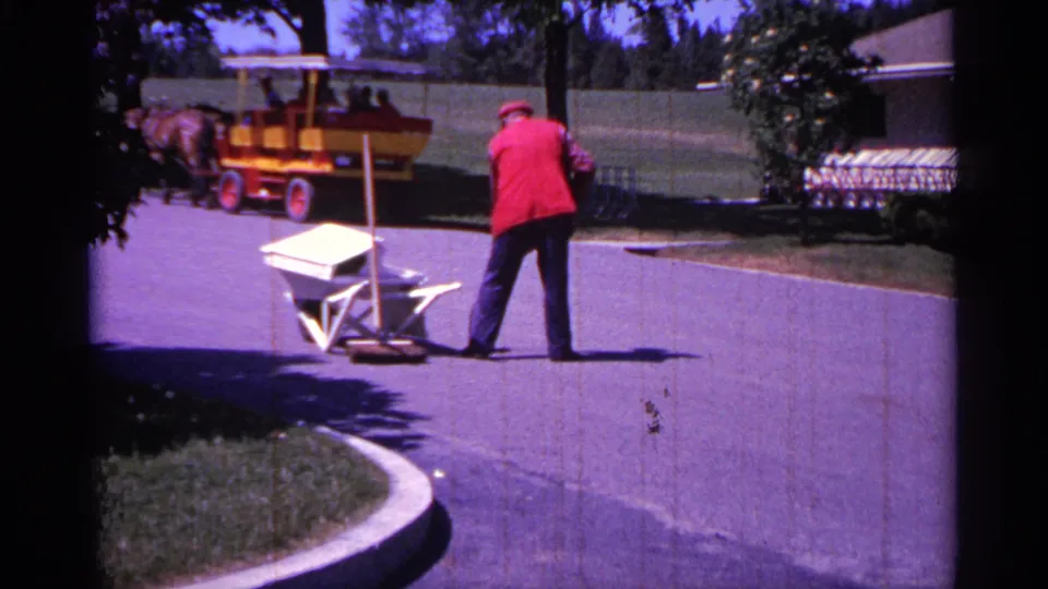 The worker stands with a broom on the paved road as the carriage and horses move away in bright 1960 Michigan home-movie footage.