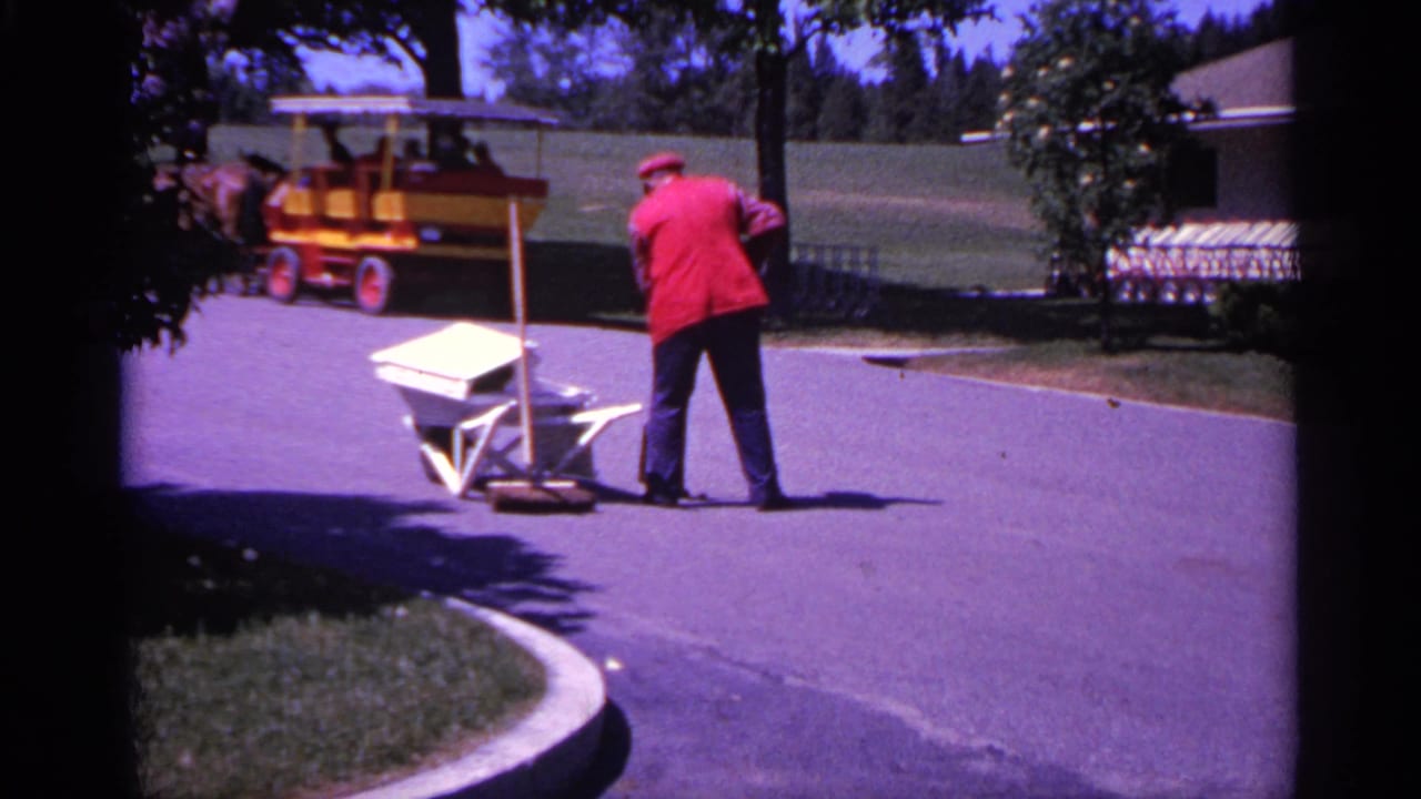 1960 Michigan 8mm frame showing a red-jacketed worker pushing a wheelbarrow along a paved park path with trees and a small tourist vehicle behind.