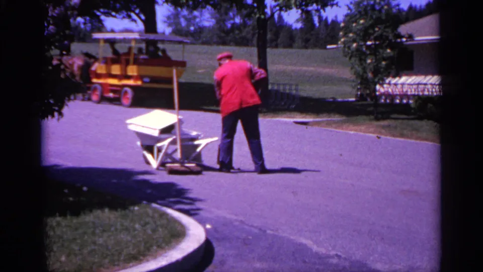 1960 Michigan 8mm frame showing a red-jacketed worker pushing a wheelbarrow along a paved park path with trees and a small tourist vehicle behind.