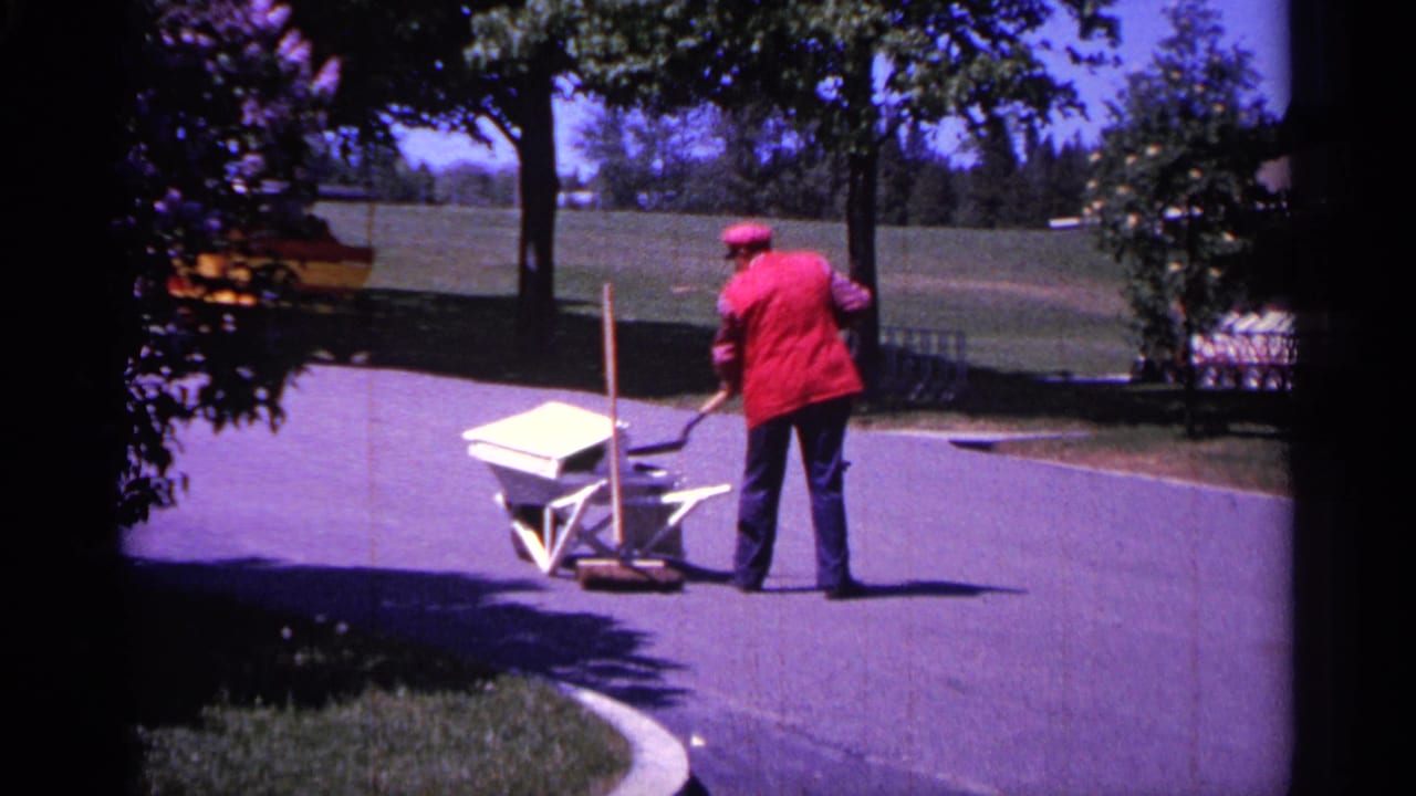 Red-jacketed worker stands with a wheelbarrow on a paved path under tree shade.
