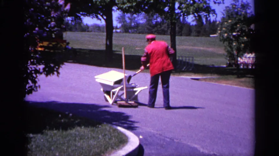 1960 Michigan 8mm frame showing the worker and wheelbarrow from behind on a curved park path, with trees and open grass in view.