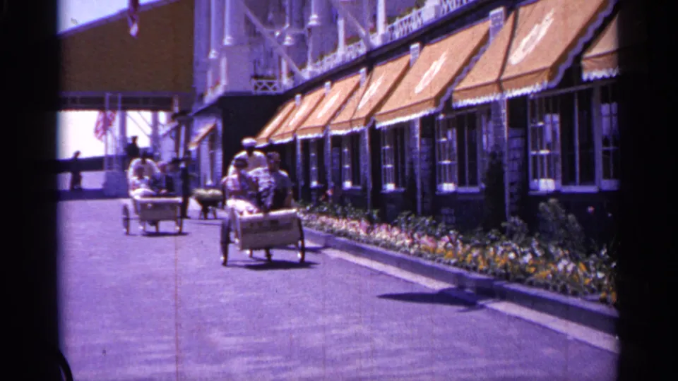 1960 Michigan 8mm frame showing seated visitors riding small carts beside a building with orange awnings and flower beds along the walkway.