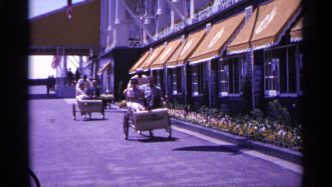1960 Michigan 8mm frame showing rickshaw-style passenger carts approaching along a sunny walkway beside orange awnings and flowers.