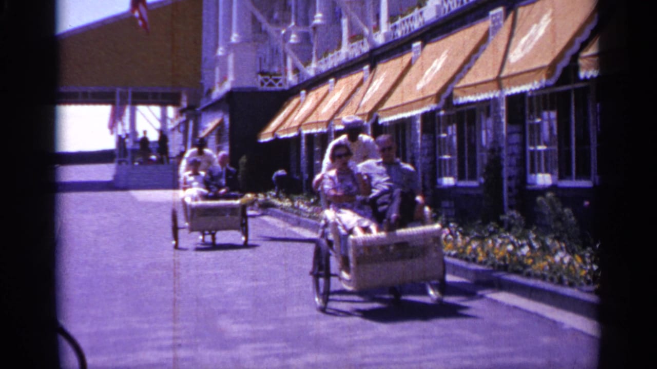 Two riders in a rickshaw-style cart pass near buildings with orange awnings and flowers.