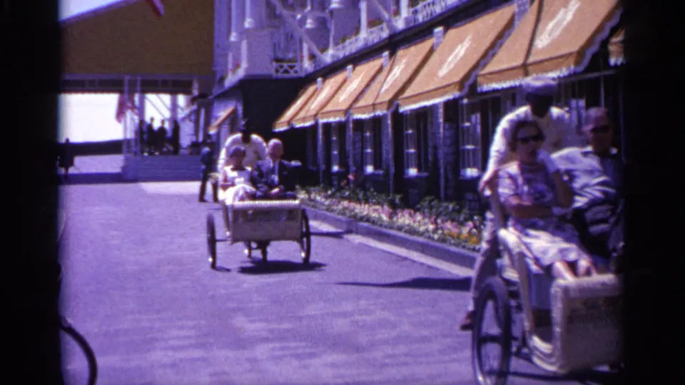 1960 Michigan home-movie frame with a rickshaw-style cart passing in the foreground and more riders moving along the landscaped building frontage.