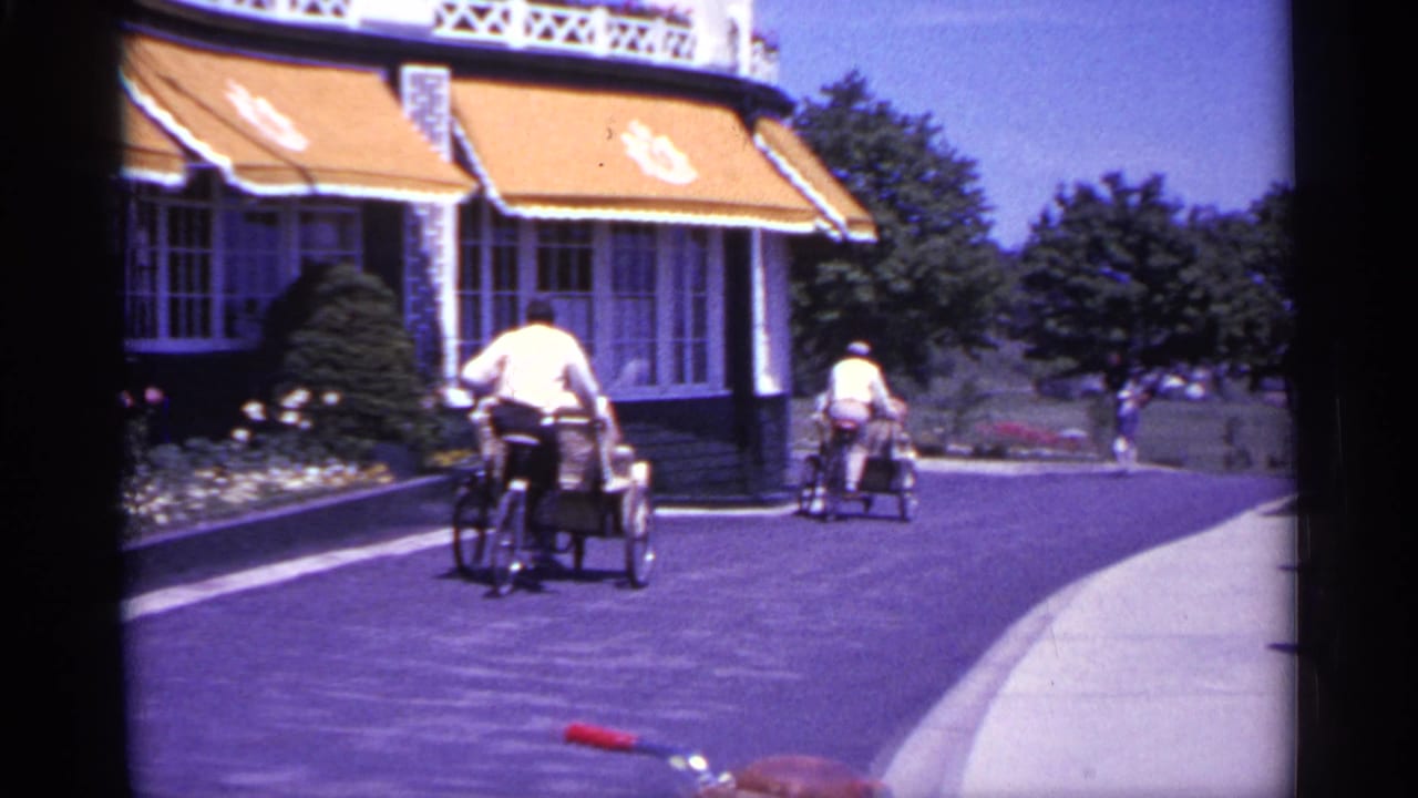 Driveway outside a large building with awnings as two riders move away from the camera.