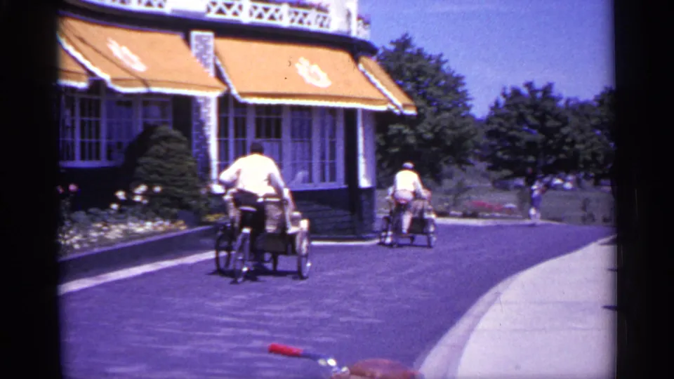 1960 Michigan home-movie view of a driveway beside a large awning-fronted building, with two riders moving away in bright daylight.