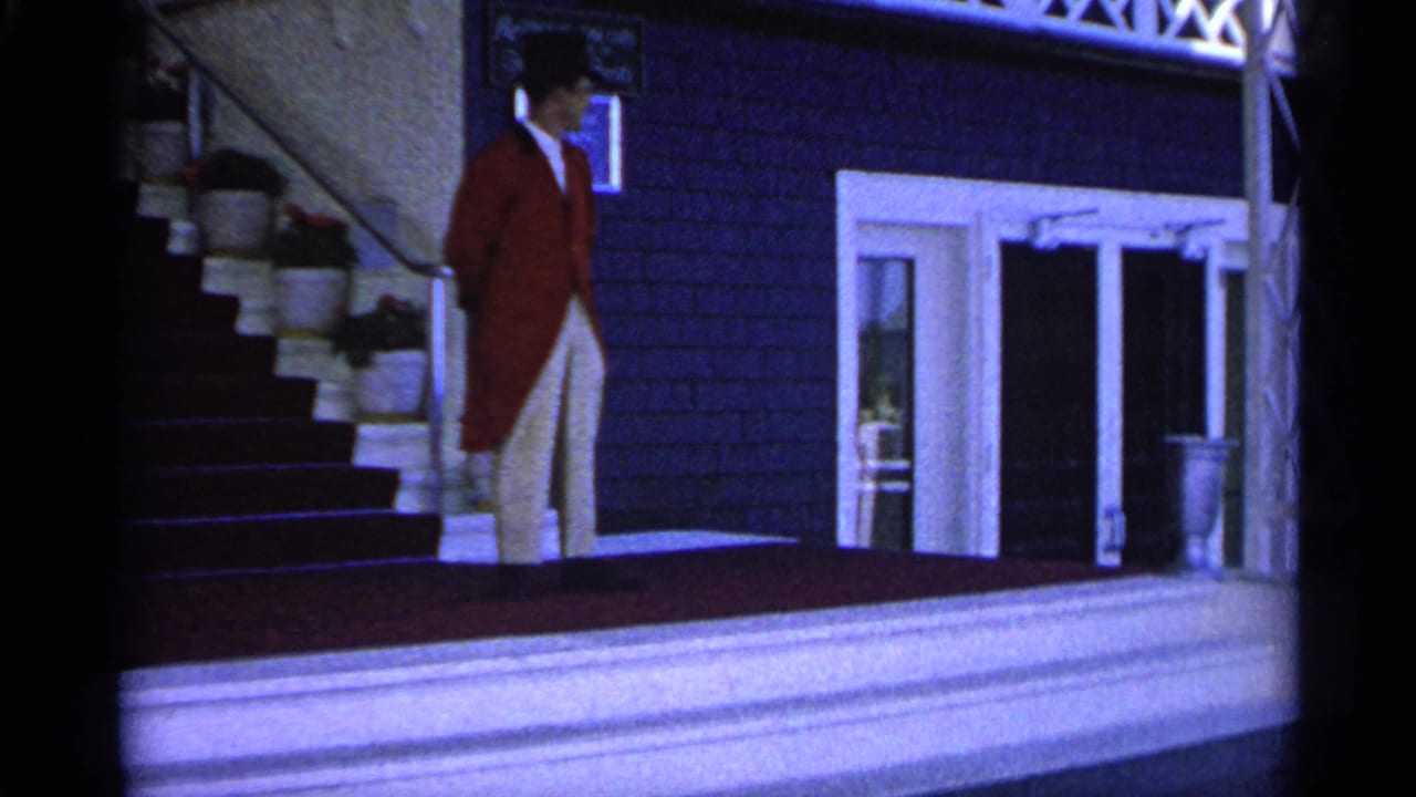 A formally dressed man in a red tailcoat, white trousers, and top hat stands on a Michigan estate porch in 1960 footage.