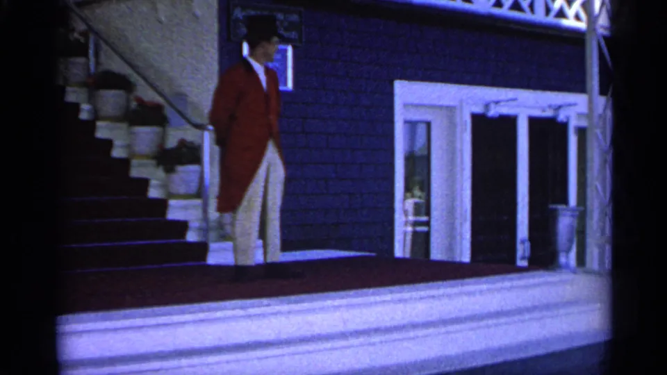 A formally dressed man in a red tailcoat, white trousers, and top hat stands on a Michigan estate porch in 1960 footage.