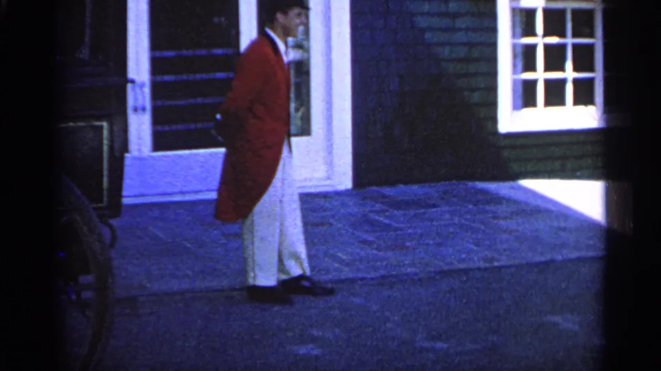 The man in red formal attire walks along the porch beside a white door and window on the Michigan estate exterior.