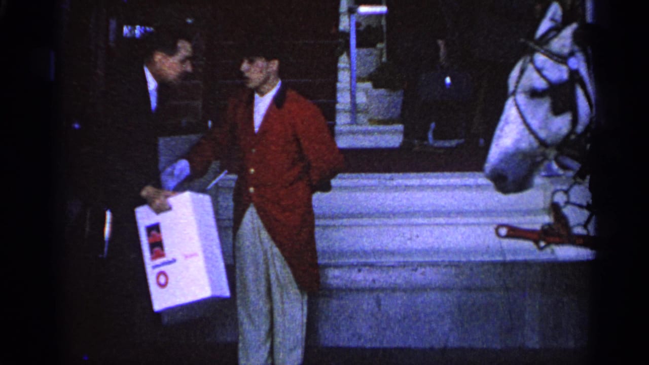 1960 Michigan 8mm frame of a suited man handing a white box to a red-coated attendant near a harnessed white horse.