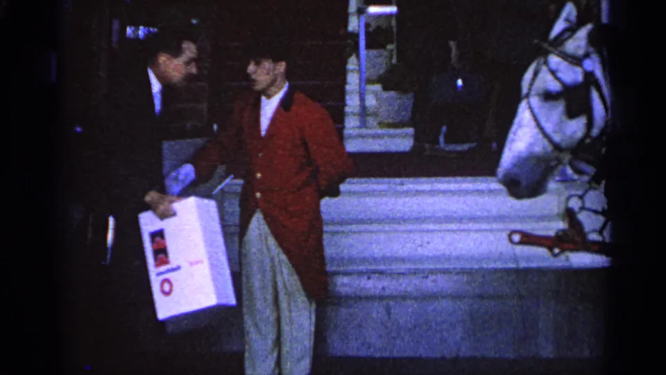 1960 Michigan 8mm frame of a suited man handing a white box to a red-coated attendant near a harnessed white horse.