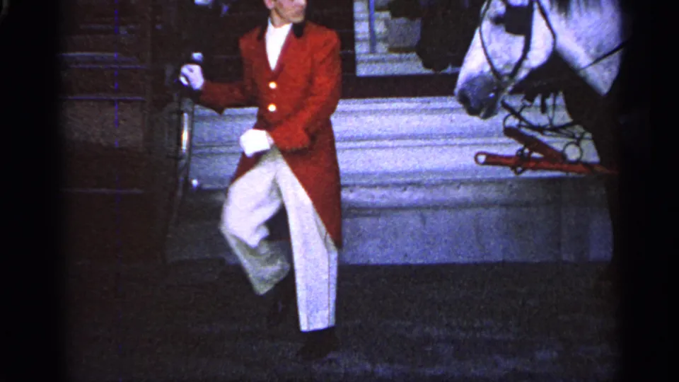 1960 Michigan 8mm frame of a red-coated, top-hatted man walking past a harnessed white horse outside a building.