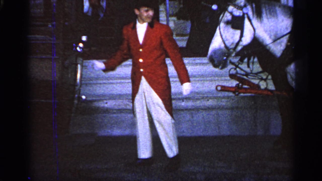 Man in red tailcoat stands beside a white horse in harness.