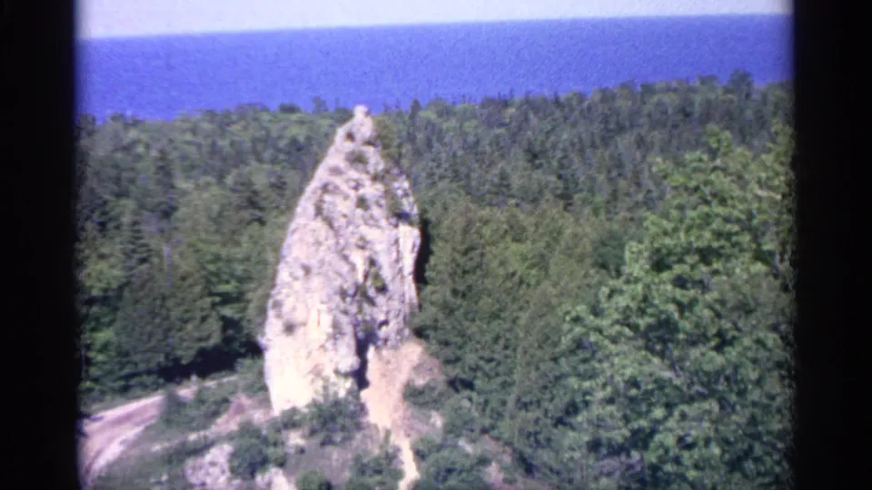 A Michigan landscape view shows a prominent rock formation above dense forest, with a blue lake in the distance.