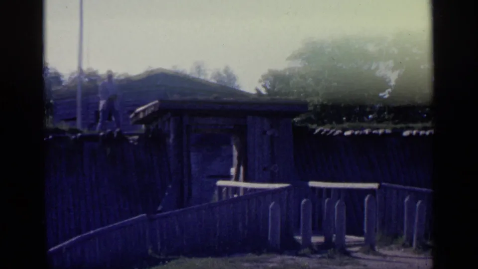 1960 Michigan 8mm frame showing a wooden fence and ramp in front of a fort-like timber structure, with trees beyond.