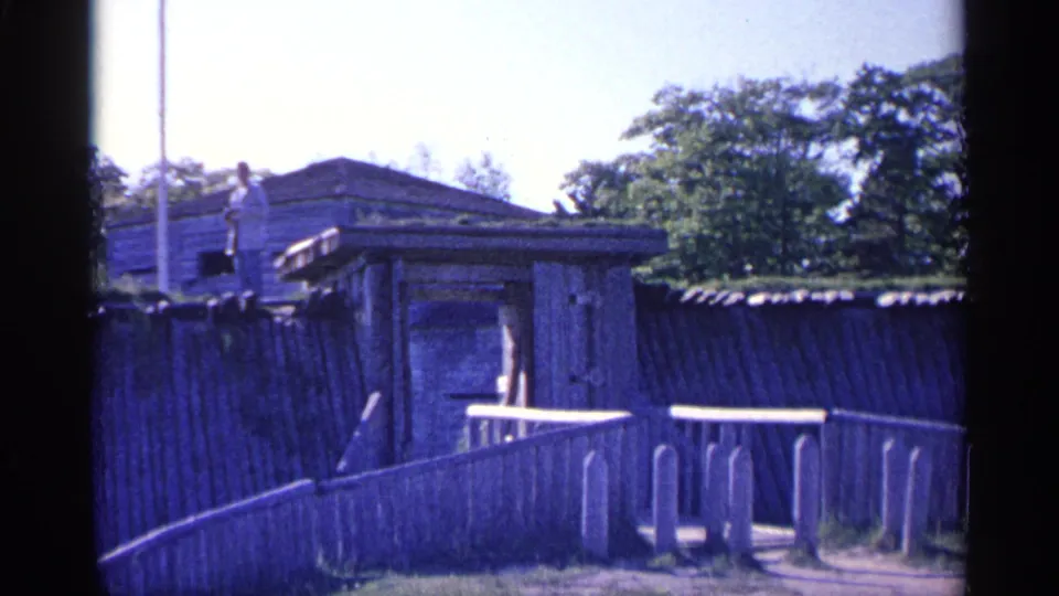 1960 Michigan home-movie frame of the fort entrance area, with a small visitor visible on the ramparts above the wooden structure.