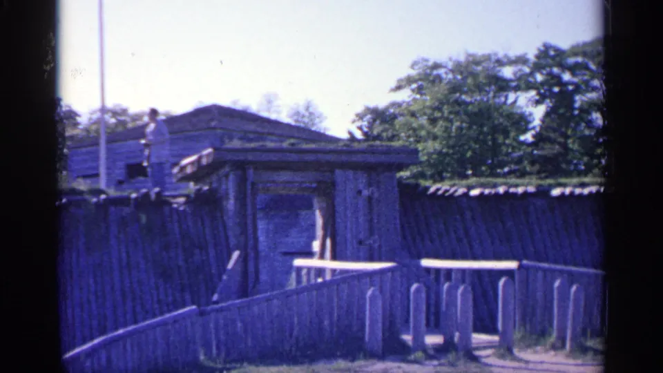 1960 Michigan home-movie frame showing the fort fence and ramp, with a distant visitor standing near the upper timber wall.
