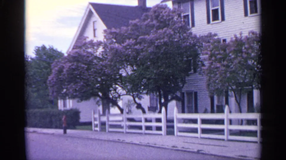 1960 Michigan 8mm frame showing a white clapboard house, white fence, sidewalk, trees, purple lilacs, and a distant visible figure.