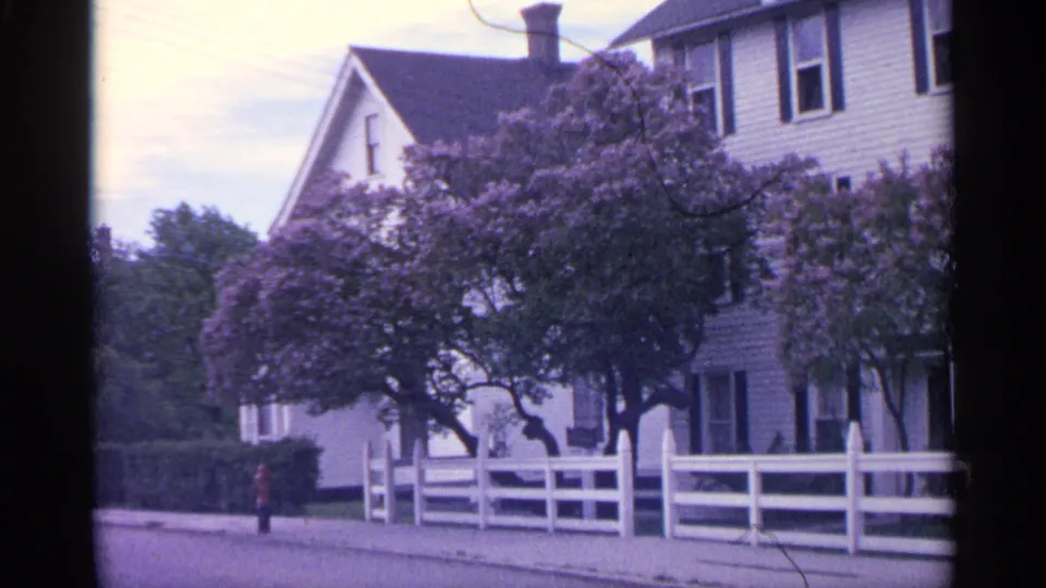 1960 Michigan home-movie frame of a white house behind a fence, with a sidewalk, mature trees, purple lilacs, and a distant visible figure.