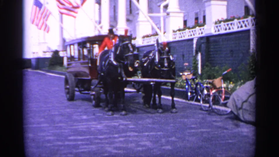 In 1960 Michigan footage, a man in a red jacket sits on a stagecoach with two dark horses near American flags, bicycles, and a white building.