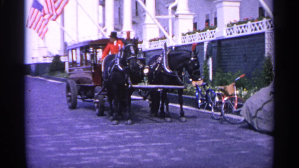 The stagecoach remains parked in the 1960 Michigan scene, with two horses, a seated driver, American flags, bicycles, and a white building visible.