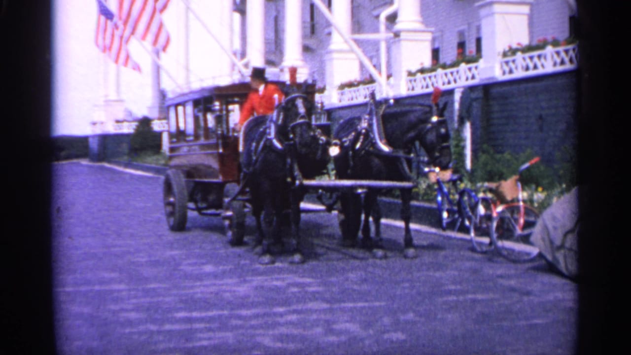 1960 Michigan 8mm frame showing a horse-drawn carriage near a white building with American flags and bicycles nearby.