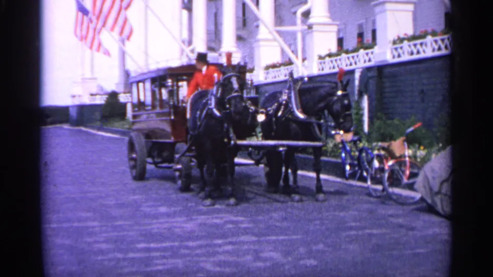 1960 Michigan 8mm frame showing a horse-drawn carriage near a white building with American flags and bicycles nearby.