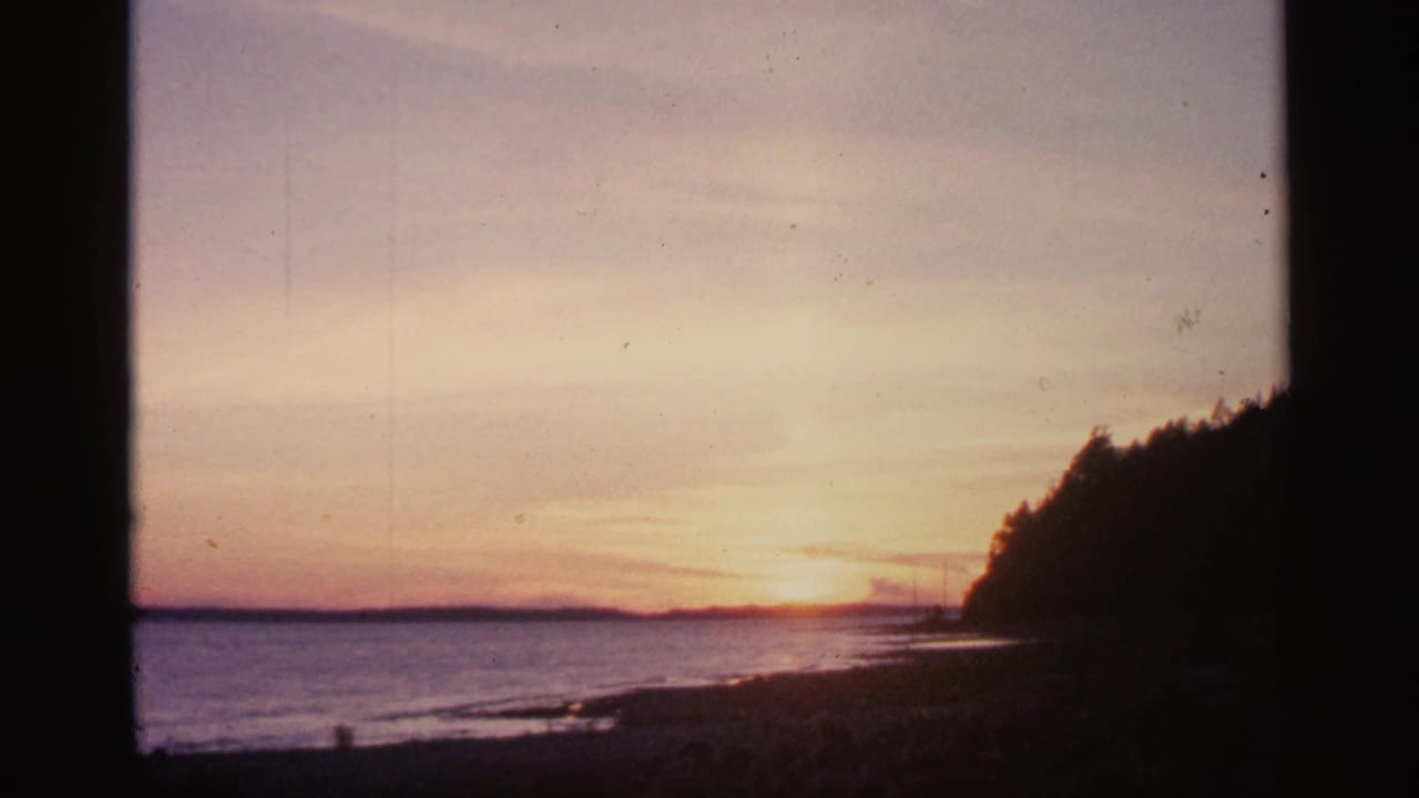 1960 Michigan 8mm frame of a warm sunset over calm water, with a silhouetted shoreline and treeline at right.