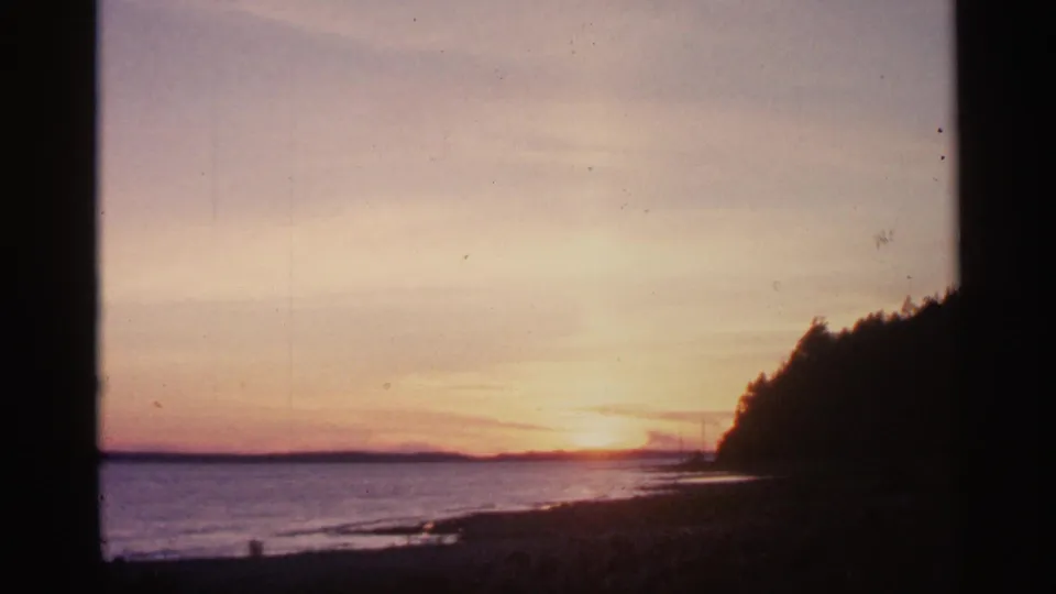 1960 Michigan 8mm frame of a warm sunset over calm water, with a silhouetted shoreline and treeline at right.