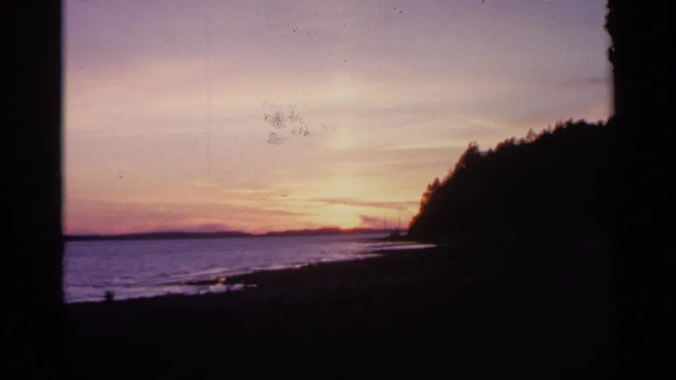 1960 Michigan 8mm frame showing sunset light over water, a dark wooded shoreline, and specks of film wear in the sky.
