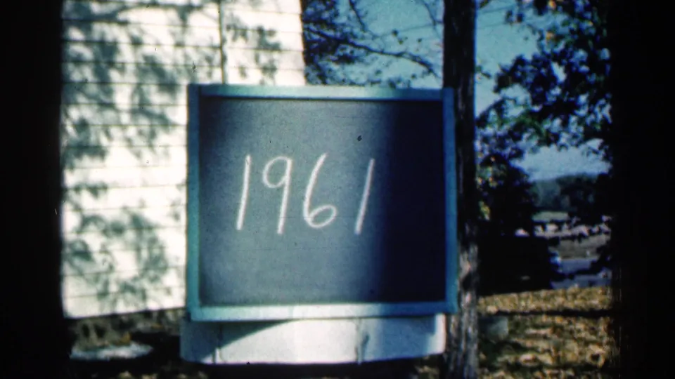 1961 Minnesota home-movie frame showing a chalkboard marked 1961 outdoors near house siding, trees, shadows, and autumn leaves.