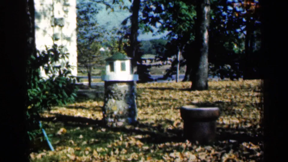 1961 Minnesota yard scene with trees, fallen leaves, a small lighthouse-style garden feature, a planter, and distant houses or structures.