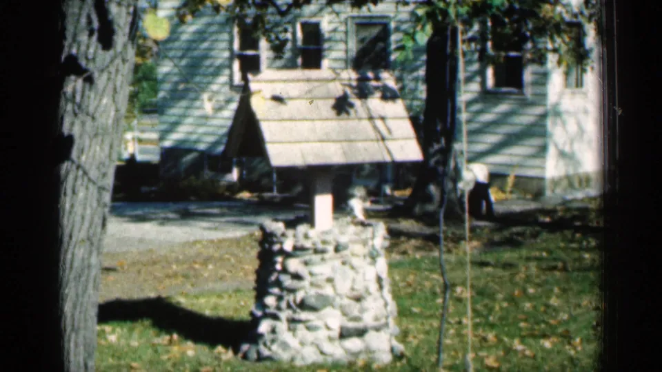 1961 Minnesota 8mm frame showing a stone wishing-well yard feature, house siding and windows, tree trunks, and a small distant person.
