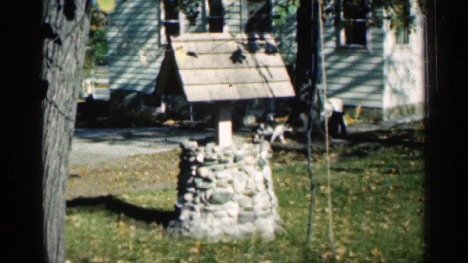 1961 Minnesota home-movie frame of a rustic stone well ornament in a residential yard, with house siding, trees, and a small person at right.