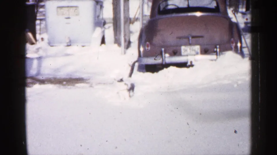 1961 Minnesota winter frame with a small black and white cat moving through snow in front of a classic car parked near a building.