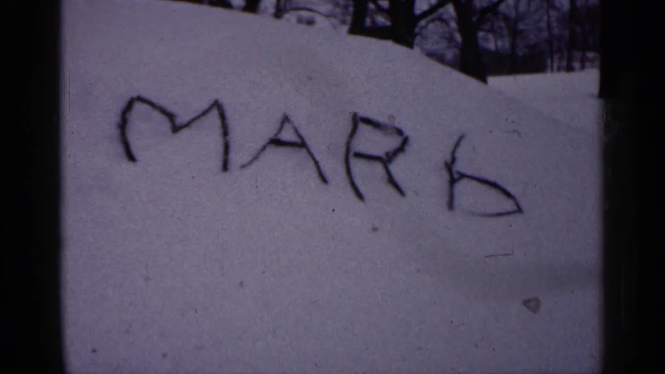 The name MARK appears written in snow in this Minnesota winter home-movie frame, with dark trees and film edges nearby.