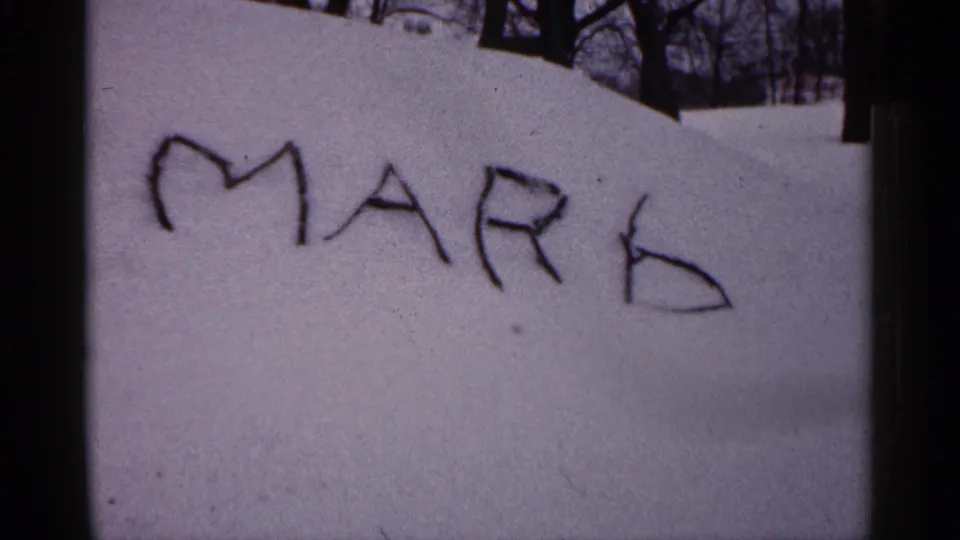 1961 Minnesota 8mm frame showing MARK written in snow on a buried car, with bare trees and a snowy yard behind it.
