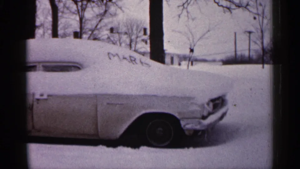 1961 Minnesota frame of a mid-century car nearly covered by snow, with MARK visible on the hood area and winter trees behind.