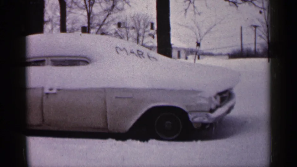 Side view of the snow-buried car in 1961 Minnesota, with bare trees, utility poles, and a quiet snowy residential background.