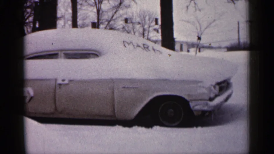 1961 Minnesota home-movie frame showing the snow-laden car from the side, with MARK on the hood and winter trees in the background.